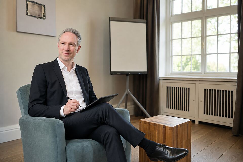 Matthias Drevs sitzt mit Stift und Schreibmappe im Sessel, im Hintergrund ein Flipchart